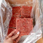 Hand holding a square raw dog food patty by RawFedK9 above a cardboard box with more raw dog food.