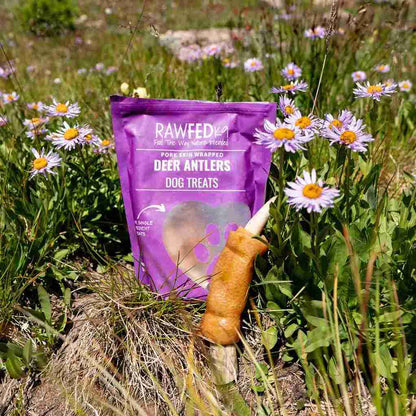 RawFedK9 pork skin–wrapped deer antler with branded bag, displayed in a natural field of blooming flowers.
