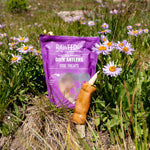 RawFedK9 pork skin–wrapped deer antler with branded bag, displayed in a natural field of blooming flowers.