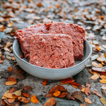 RawFedK9 PMR patties served in a blue dog bowl placed on the ground among colorful fall leaves, showcasing a balanced raw meal in a seasonal outdoor setting.